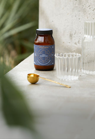 An amber glass bottle of Ancient and Brave True Hydration on a stone ledge beside a ribbed clear glass cup and a clear glass bottle, with a small gold spoon in front. Blurred green leaves are in the background.